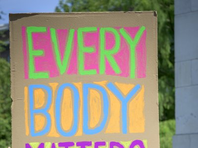 A man stands in front of a colorful, large cardboard sign in vibrant pink, blue, and purple letters.