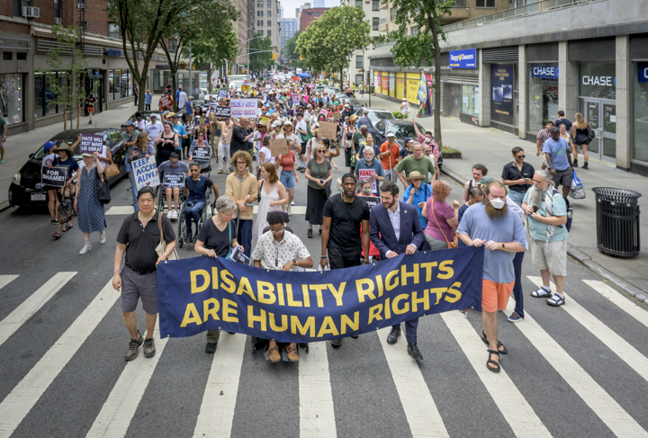 A large crowd of marchers walks down a city street behind a dark blue banner.