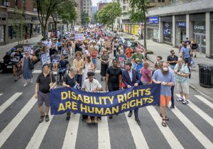 A large crowd of marchers walks down a city street behind a dark blue banner.