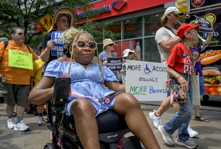 A woman in a motorized wheelchair leads a group of marchers while holding a sign about accessibility.