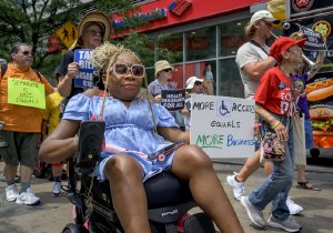 A woman in a motorized wheelchair leads a group of marchers while holding a sign about accessibility.