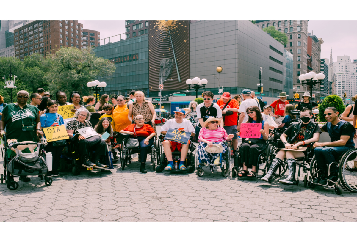 Group of disability rights activists sitting in wheelchairs and standing outdoors holding protest signs.