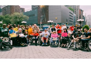 Group of disability rights activists sitting in wheelchairs and standing outdoors holding protest signs.