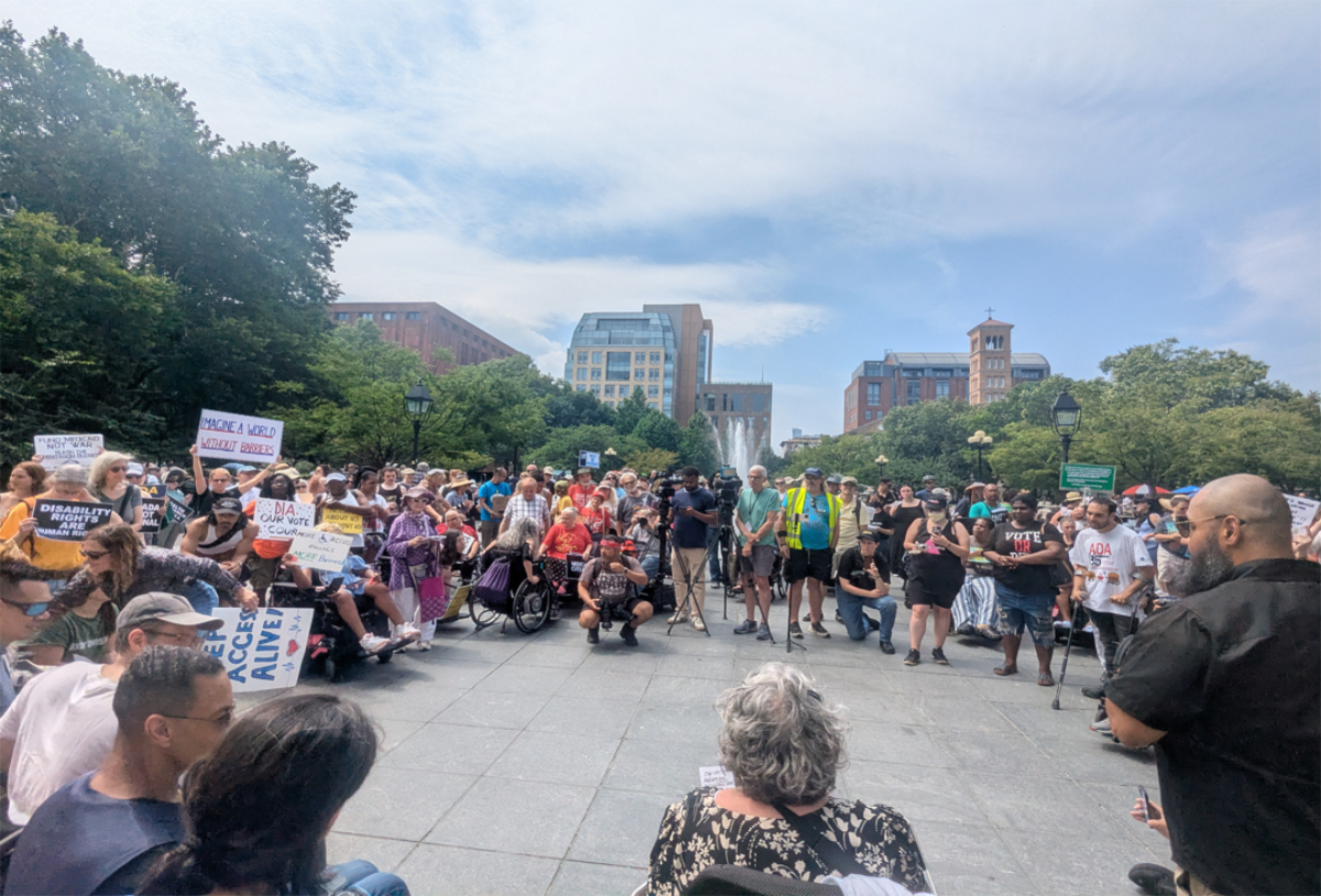Crowd of people gathered in a park plaza for a disability rights rally holding protest signs.