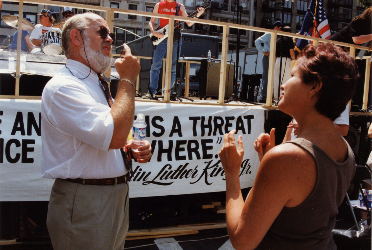 Man with a beard and sunglasses signing to a woman outdoors beside a rally banner.