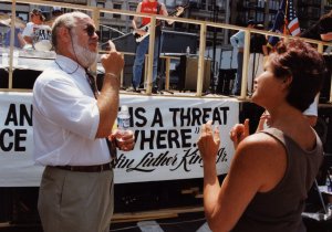 Man with a beard and sunglasses signing to a woman outdoors beside a rally banner.
