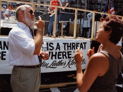 Man with a beard and sunglasses signing to a woman outdoors beside a rally banner.