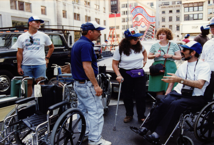 A group of people stands on a city street surrounded by a collection of wheelchairs.