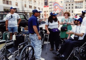 A group of people stands on a city street surrounded by a collection of wheelchairs.