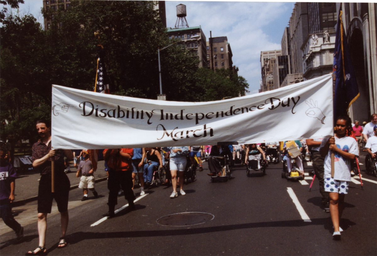 Two people holding a wide white banner as a crowd marches down a city street.