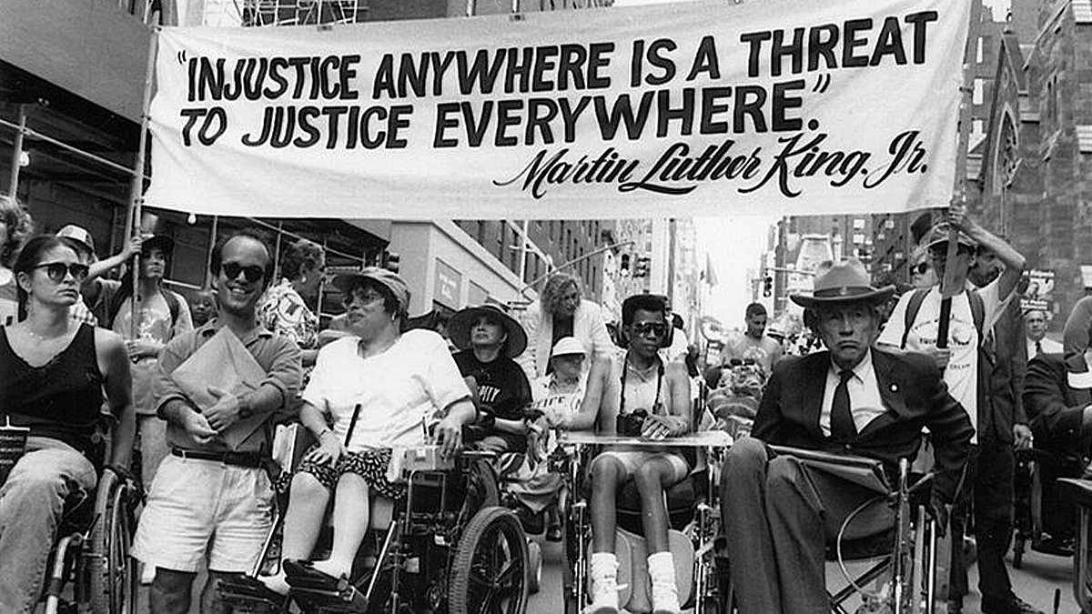 Six disability rights activists stand side by side in NYC, holding a large banner featuring a Martin Luther King Jr. quote about justice.
