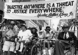 Six disability rights activists stand side by side in NYC, holding a large banner featuring a Martin Luther King Jr. quote about justice.