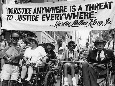 Six disability rights activists stand side by side in NYC, holding a large banner featuring a Martin Luther King Jr. quote about justice.