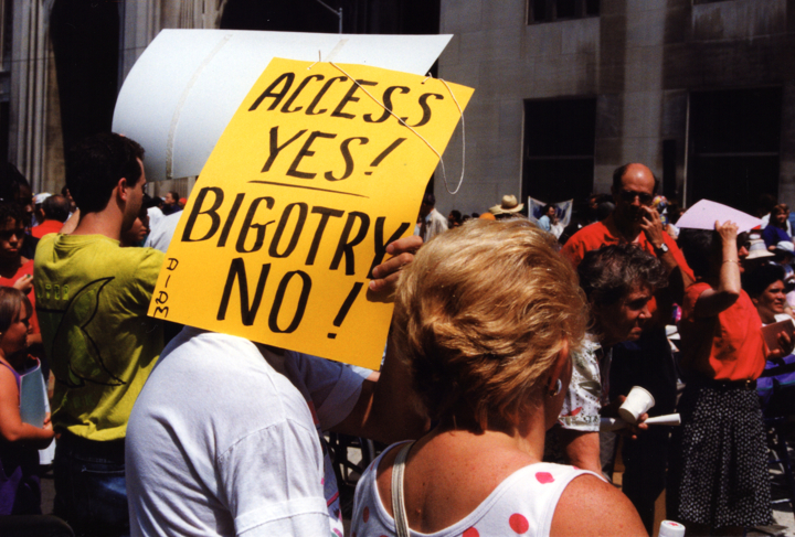 A protester in a crowd holds up a bright yellow sign, hiding their face.