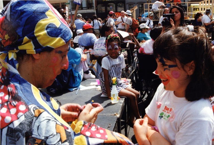 Chidren have their faces painted like clowns by an adult with clown makeup