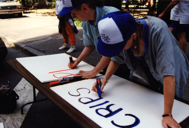 Two people wearing matching baseball caps lean over a table outdoors, writing on large poster boards with markers.