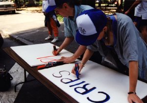 Two people wearing matching baseball caps lean over a table outdoors, writing on large poster boards with markers.