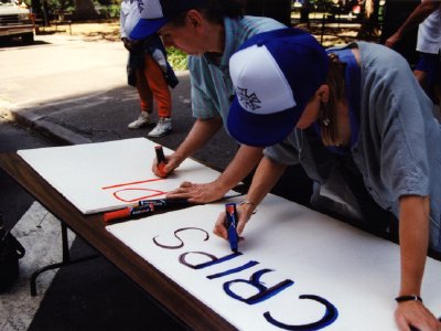Two people wearing matching baseball caps lean over a table outdoors, writing on large poster boards with markers.