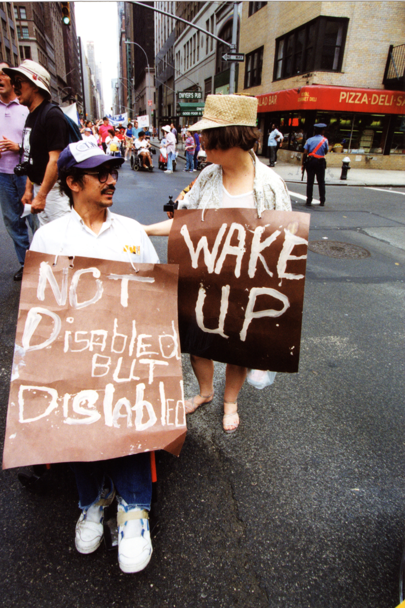 Two disability rights marchers hold large, handwritten cardboard signs on a city street