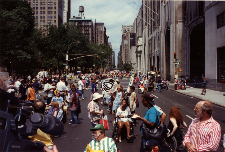 A blue police car leads a large group of marchers down a major city street.