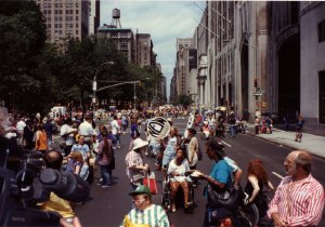 A blue police car leads a large group of marchers down a major city street.