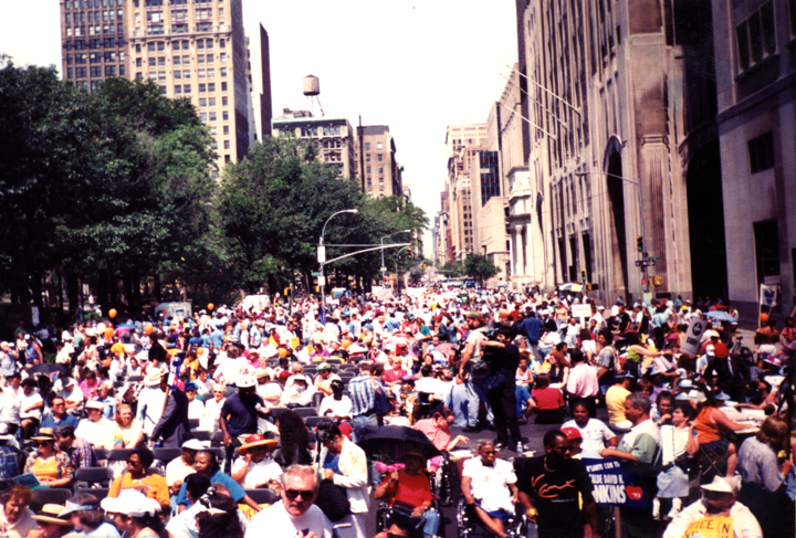 Protesters and wheelchair users gather on a wide city street during a demonstration.