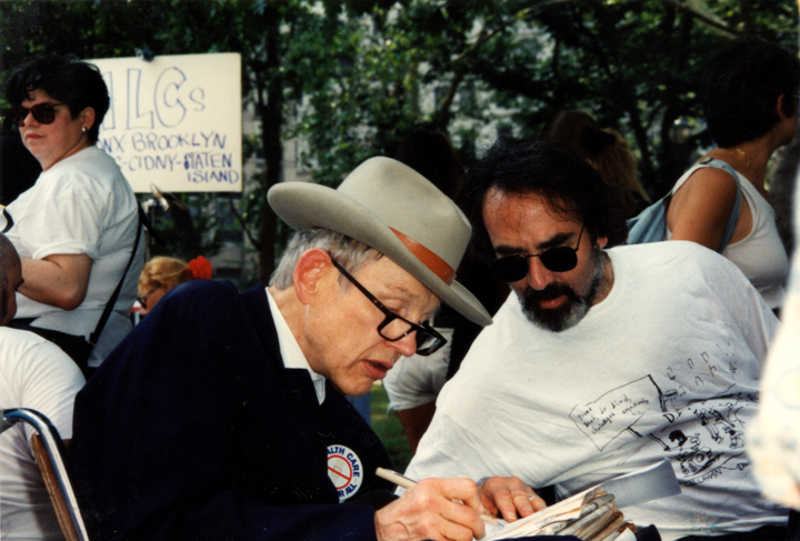 Two men seated outdoors conferring closely while one signs or writes on paperwork at an event.