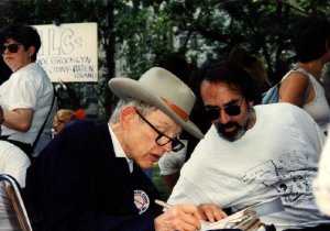 Two men seated outdoors conferring closely while one signs or writes on paperwork at an event.