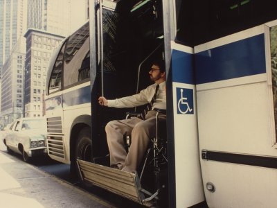 A man in a wheelchair exits a bus from a lift at the back of the bus