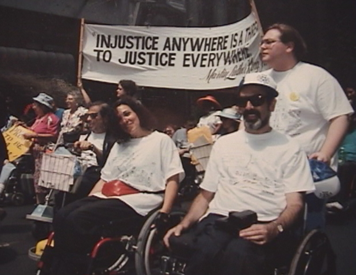 Two wheelchair users smiling together in a march under a banner quoting Martin Luther King Jr.