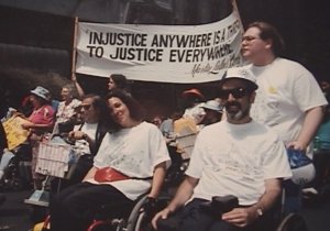 Two wheelchair users smiling together in a march under a banner quoting Martin Luther King Jr.