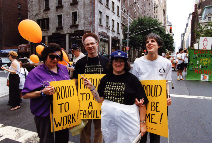 Four demonstrators stand together on a city street holding yellow signs and orange balloons.