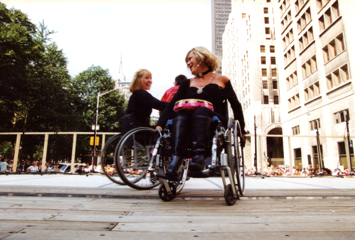 Two women in wheelchairs dance together on a wide, wooden outdoor stage during a rally.