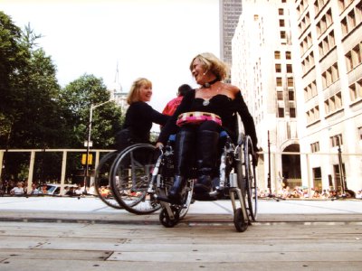 Two women in wheelchairs dance together on a wide, wooden outdoor stage during a rally.