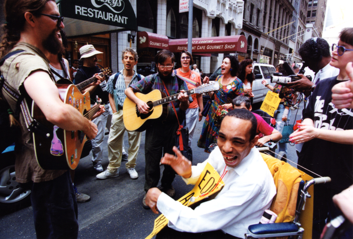 Musicians play acoustic guitars on a city street while surrounding people sing along.