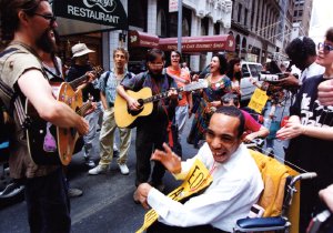 Musicians play acoustic guitars on a city street while surrounding people sing along.