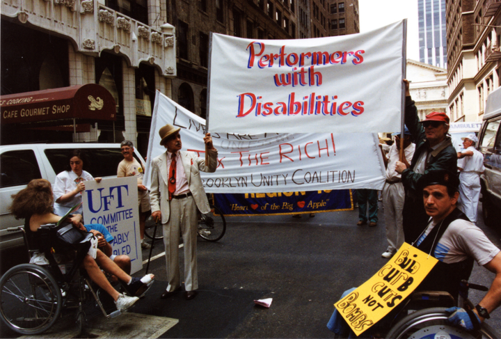 Marchers walk on a New York City street carrying large banners, including one for "Performers with Disabilities".