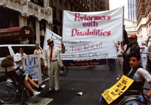 Marchers walk on a New York City street carrying large banners, including one for "Performers with Disabilities".