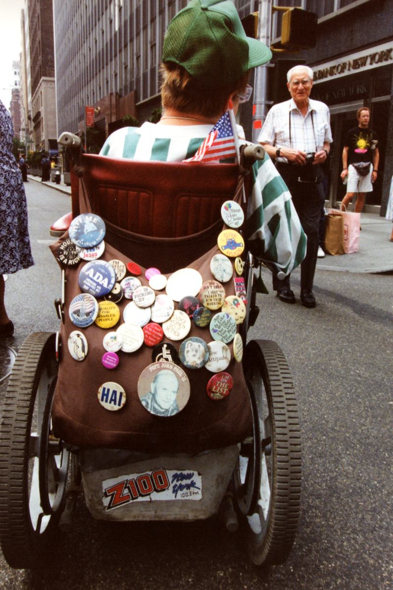 Back of a wheelchair covered in advocacy buttons, featuring an ADA badge and a small American flag.